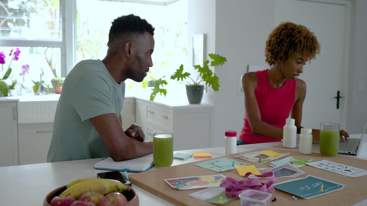 African-American duo woman picking white bottle arranging mockups using laptop in kitchen packaging