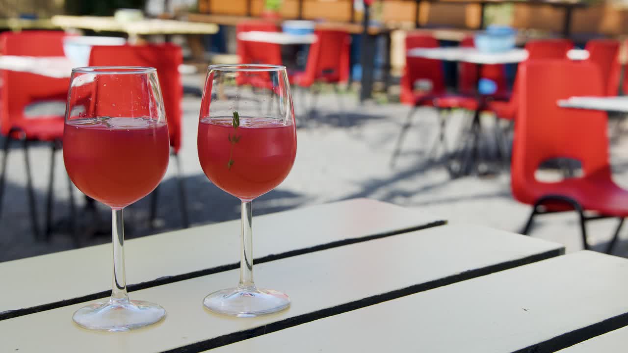 Hand lifts red cocktail from white table at sunny outdoor café with red chairs
