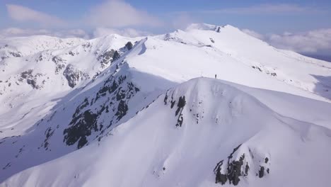 Wide-aerial-view-of-lone-climber-standing-on-snow-covered-mountain-peak-with-view-of-landscape-below