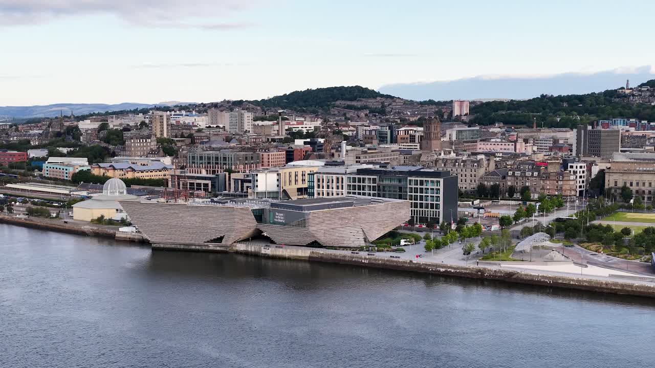 Drone pans over contemporary waterfront museum, cityscape, and river under soft daylight in Dundee