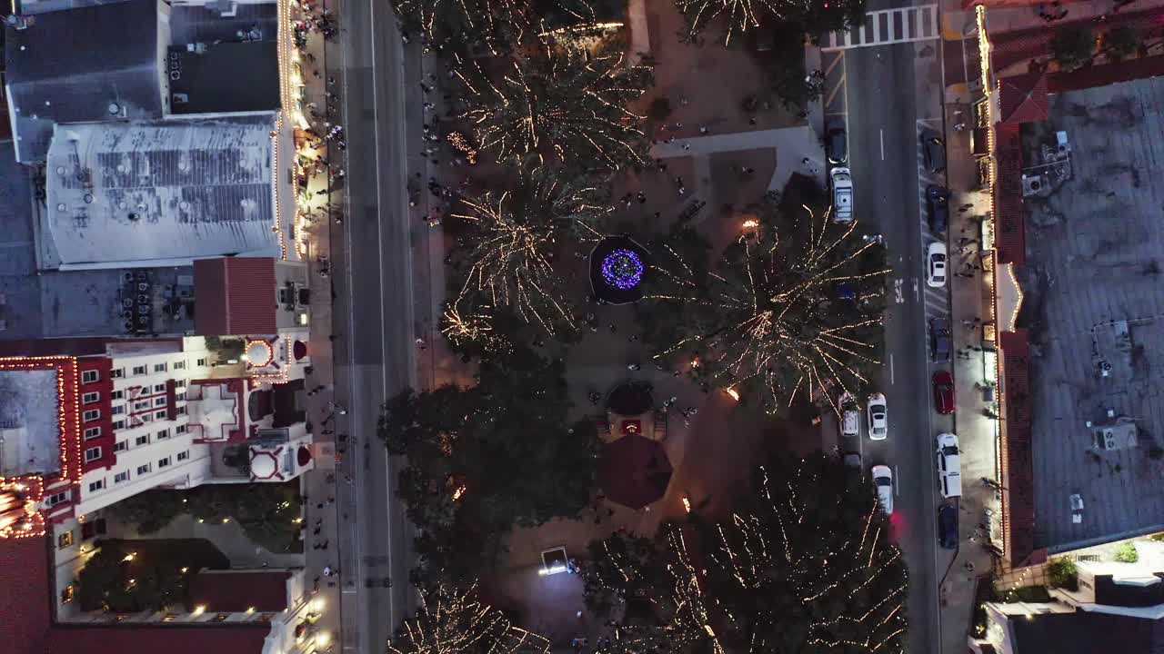 Parco del centro decorato con luminose luci natalizie, circondato da strade cittadine, veicoli ed edifici di notte, creando un'atmosfera urbana festosa in questa vista aerea.