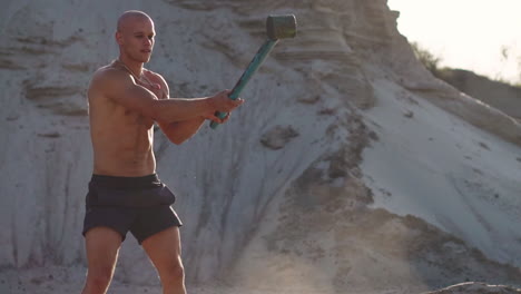 bald man strongman hits a hammer on a huge wheel in the sandy mountains in slow motion. strength and endurance training for wrestlers.