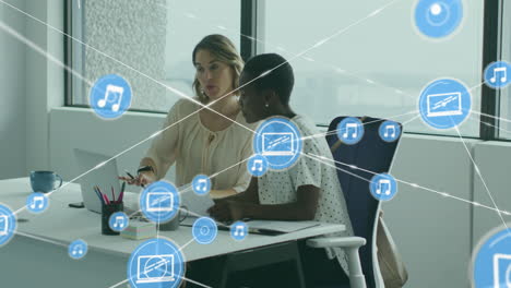 Animation-of-connected-icons-over-diverse-female-coworkers-discussing-reports-on-laptop