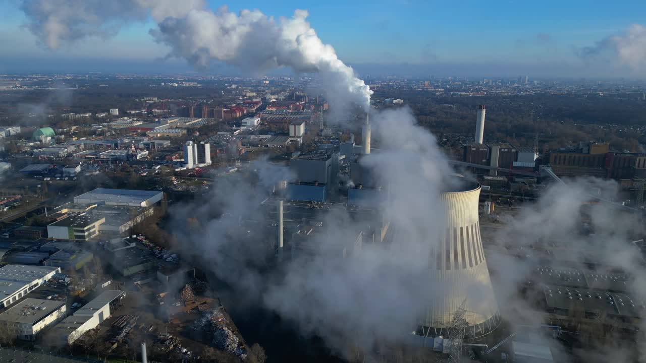 Aerial view of a thermal power plant emitting smoke on a sunny day, with a river and a city in the background. Unique aerial view flight fly reverse drone