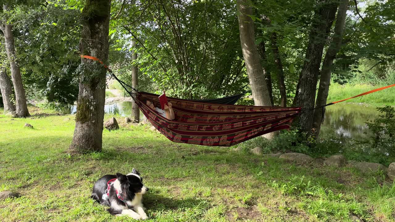 una chica asiática leyendo un libro en una hamaca junto al río, con su perro