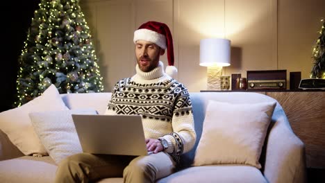 Portrait-Of-Happy-Young-Man-In-Santa-Hat-Sitting-In-Decorated-Room-Near-Glowing-Xmas-Tree-Speaking-On-Video-Chat-On-Laptop