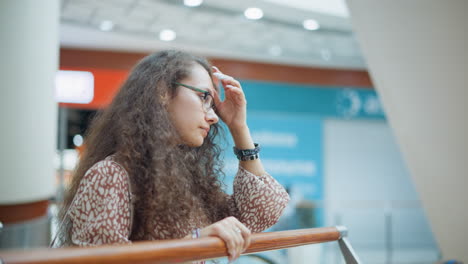 woman stands thoughtfully with hand on railing, adjusting her hair and appearing slightly tired, background shows another person coming up from an escalator