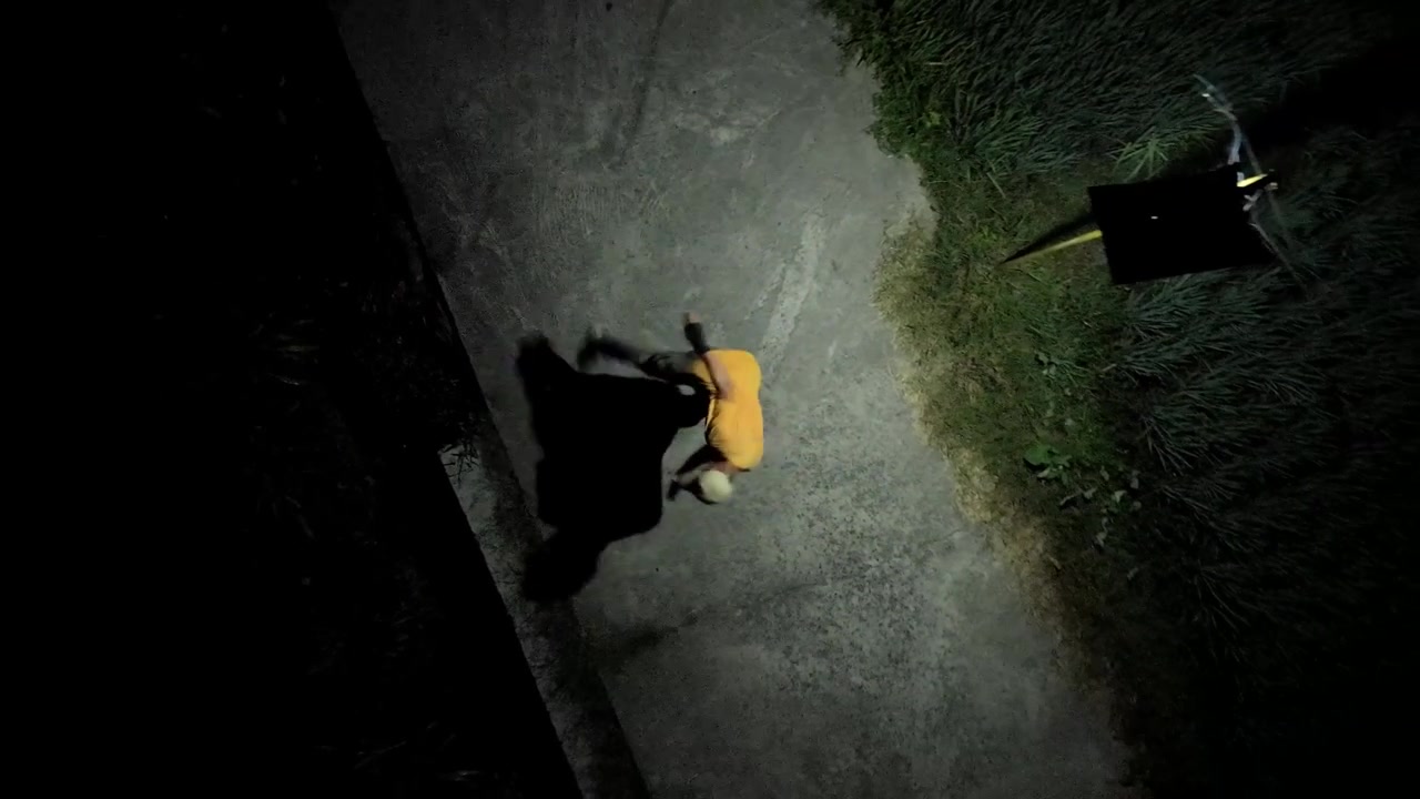 Drone in slow motion che sale e ruota rivelando una vista aerea dall'alto di un uomo con un berretto che fa shadow boxing di notte. Si allena nelle arti marziali su un sentiero esterno sotto un'illuminazione artificiale drammatica
