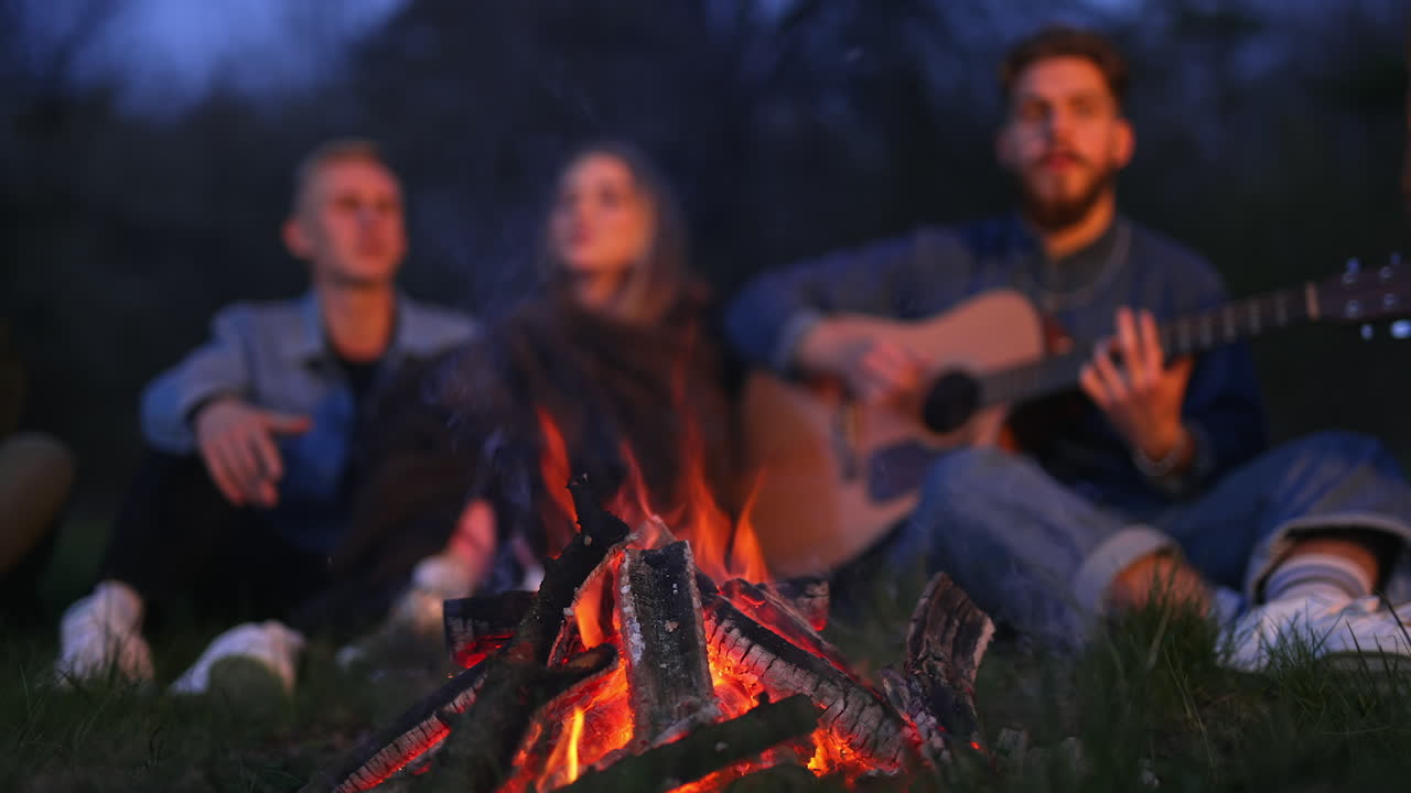 Campfire burning brightly near the forest in the evening. Friendly company warming themselves near fire in blur at the backdrop.
