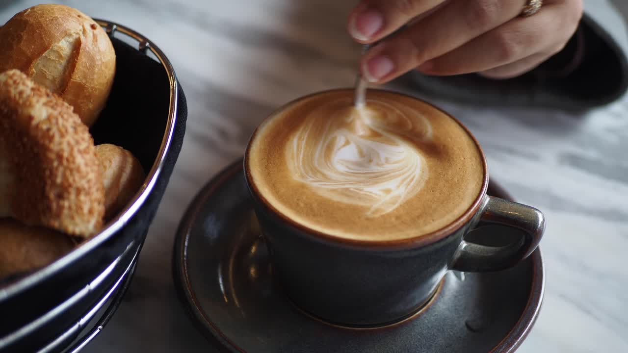 la mano de la mujer agitando una taza de arte latte con una cuchara, una canasta de rollos al lado