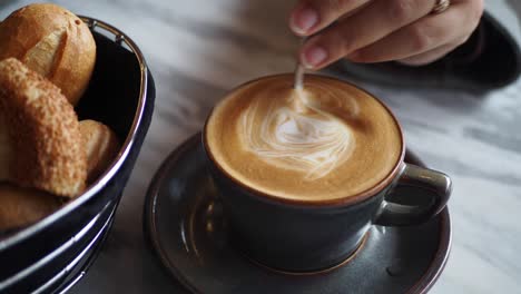 woman's hand stirring a cup of latte art with a spoon, a basket of rolls to the side