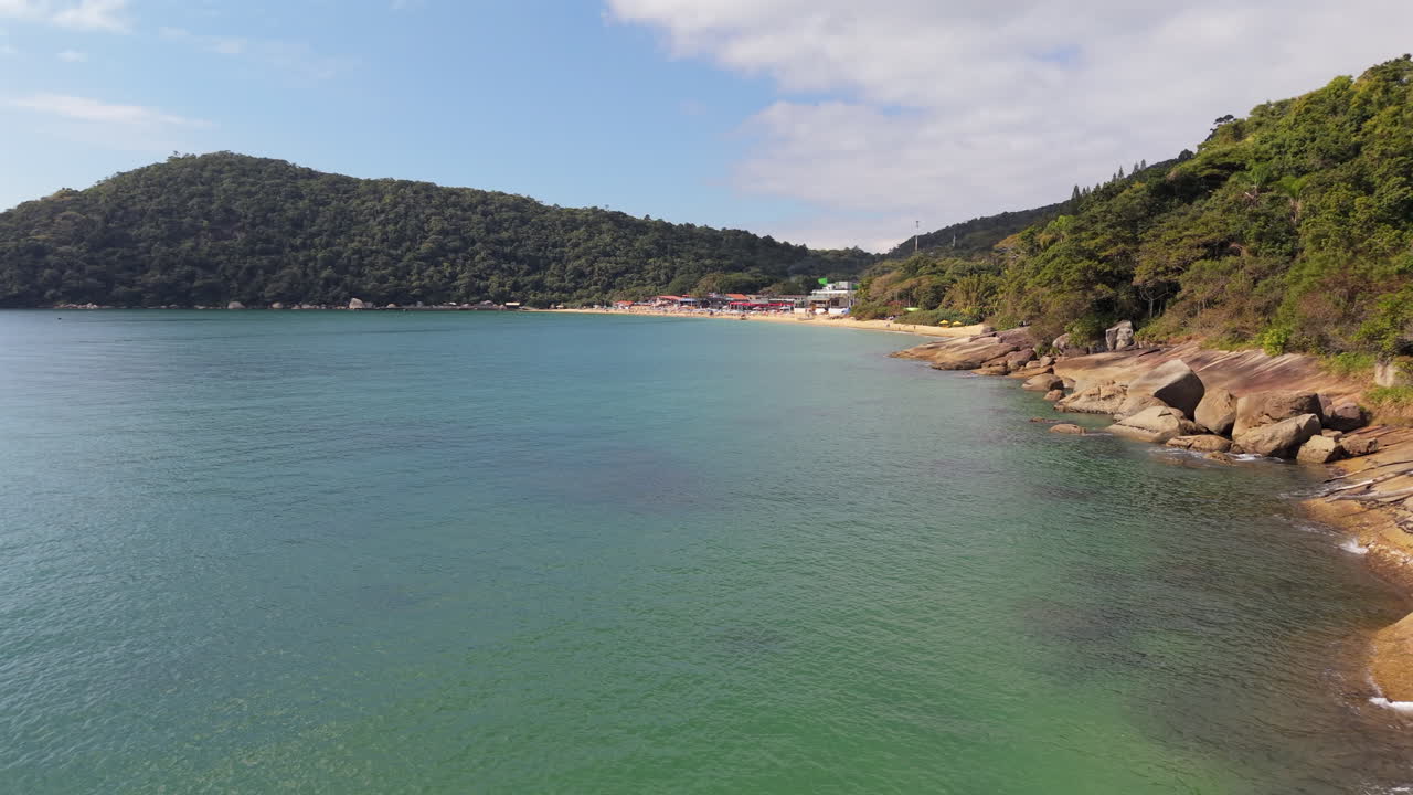 A stunning aerial approaching over the calm, clear water towards the beautiful Praia das Laranjeiras, a tropical beach nestled in a cove surrounded by lush green hills in Brazil