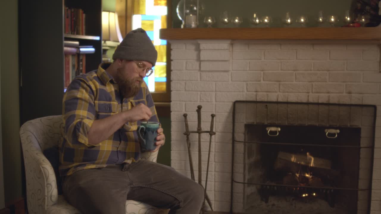 A man in plaid sits by the fireplace in a cozy fall living room setting, stirring and drinking tea. The vintage fireplace with logs on fire warms the living room. Shot in 60 frames for slow motion
