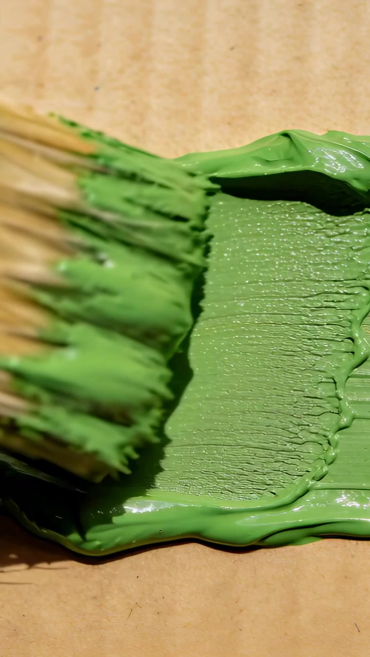 Video verticale: Le setole di un pennello premono e trascinano vernice verde su un cartone su un tavolo per formare una striscia.