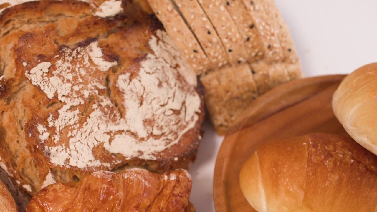 Close-up overhead shot of sourdough, croissants, and sliced whole wheat bread on white background.