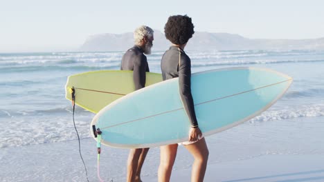 Happy african american couple walking with surfboards on sunny beach