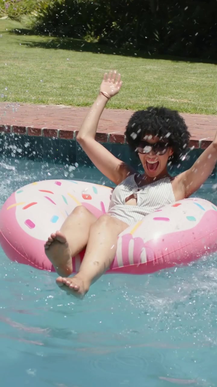 Vertical video: Sitting woman on pink donut at poolside in swimsuit and shades splashing, relaxing