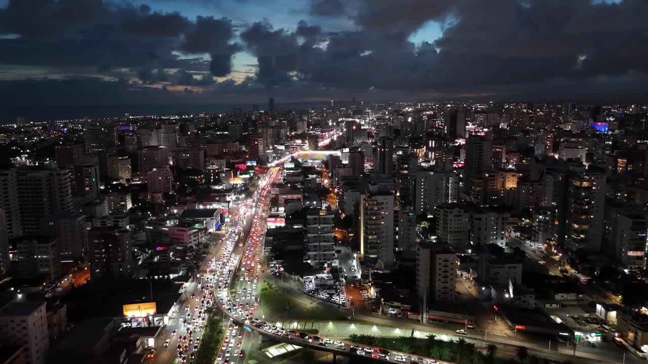 imágenes aéreas espectaculares de santo domingo por la noche, mostrando luces vibrantes de la ciudad, tráfico bullicioso, edificios altísimos y nubes a la deriva sobre el horizonte en un panorama urbano dinámico.