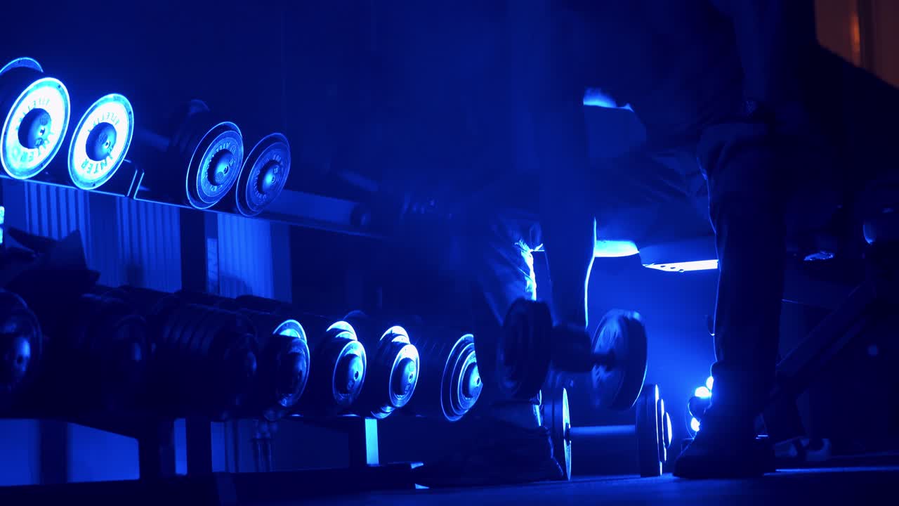 Athletic man working out in gym doing exercises with dumbbells, strong male naked torso abs. Blue dark light filter. Selective focus.