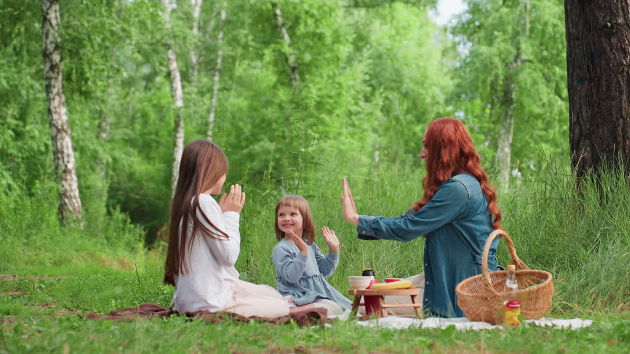 키 큰 나무와 따뜻한 햇빛이 내리쬐는 야외 피크닉 매트에 엄마와 두 딸이 앉아 함께 웃으며 눈을 감고 손을 잡고 있는 즐거운 순간