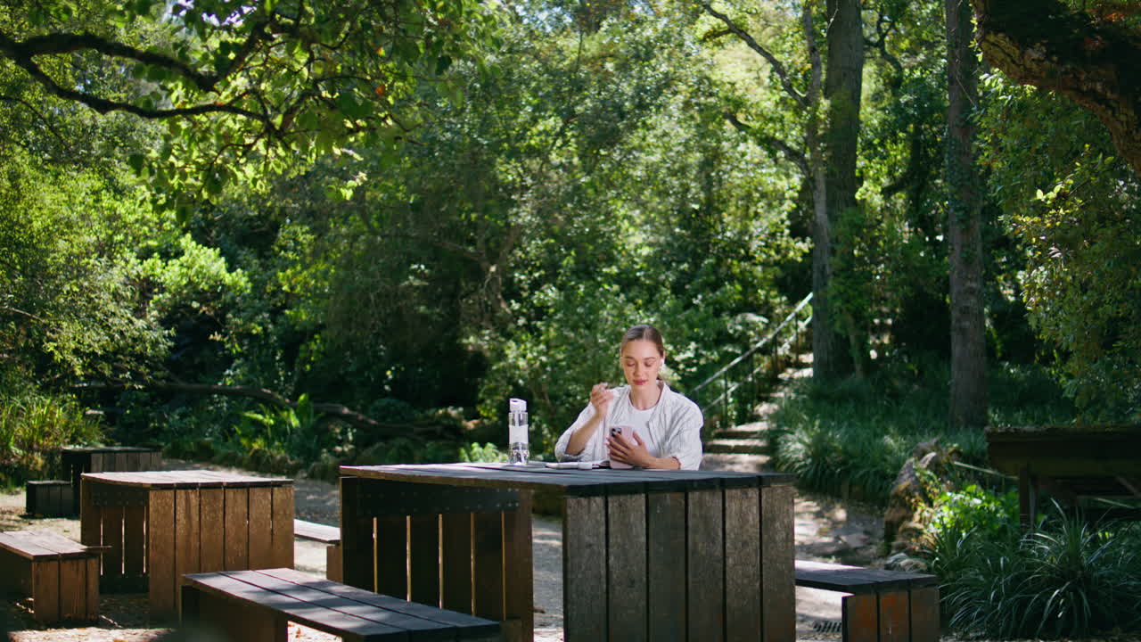 여자 프리랜서가 스마트폰을 사용하여 온라인 회의를 하고 숲의 테이블에 앉아 있습니다.