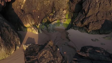 Water-spilling-onto-sandy-beach-from-cliff-face-at-evening-sunset-aerial-pull-back-shot