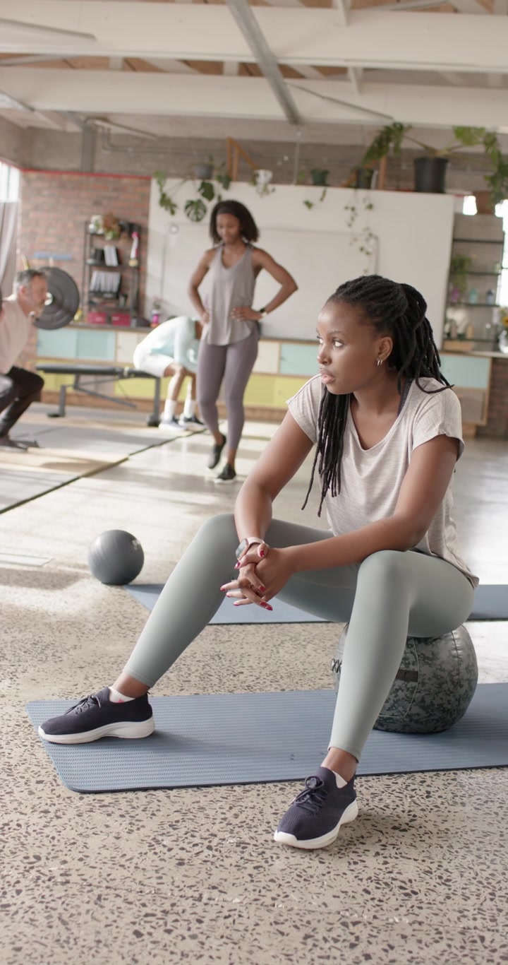 Vertical video: African American woman sitting on ball in gym resting between sets as peers moving