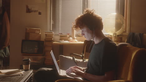joven enfocado en auriculares escribiendo en una computadora portátil en una habitación iluminada por el sol