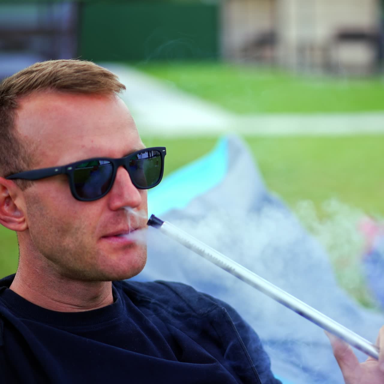 Handsome man in black clothes and sunglasses relaxing in bean bag chair smoking hookah. Male working on his laptop outdoors in a relaxed atmosphere