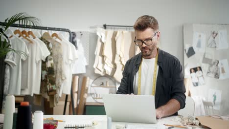 man tailor in glasses sitting at the table with a sewing machine using laptop and looks at camera