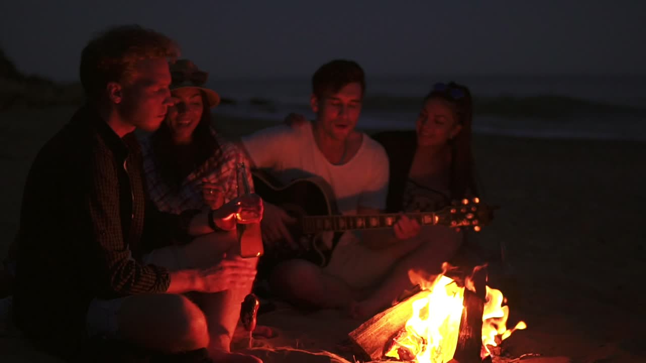 picnic de jóvenes con hoguera en la playa por la noche. amigos alegres cantando canciones y tocando la guitarra. cámara lenta