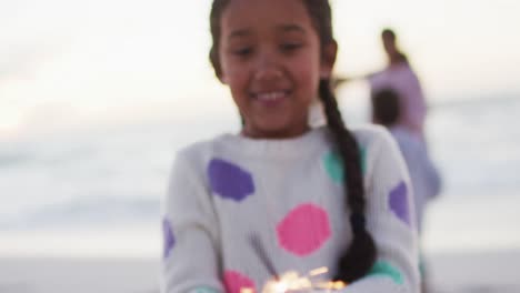 portrait of happy hispanic girl playing with sparklers on beach at sunset