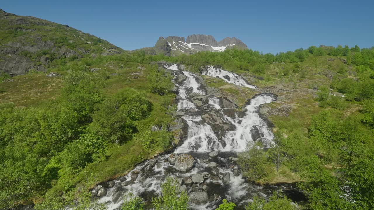 Splendida cascata di Sørvågen che scende attraverso una vegetazione lussureggiante con montagne di granito sullo sfondo, sotto un cielo azzurro e limpido, nelle Lofoten, Norvegia, lungo il sentiero escursionistico di Munkebu