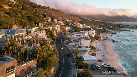 cars-drive-on-road-above-Clifton-Beach-in-Cape-Town,-South-Africa-at-sunset,-aerial