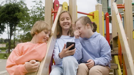 close-up view of a little girl with down syndrome and her friends laughing and watching a video on smartphone in the park on a windy day