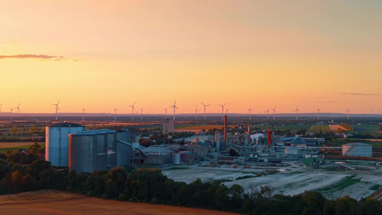 Instalação industrial em silhueta contra o pôr do sol de um parque eólico. Silos de grãos e uma chaminé alta erguem-se diante de fileiras de turbinas sob um céu noturno laranja e rosa.
