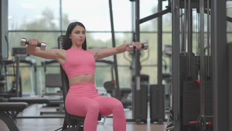 A-brunette-woman-in-a-pink-suit-raises-dumbbells-to-the-sides-while-training-her-shoulders-in-the-gym.-Seated-Shoulder-and-Arm-Bench-Exercise