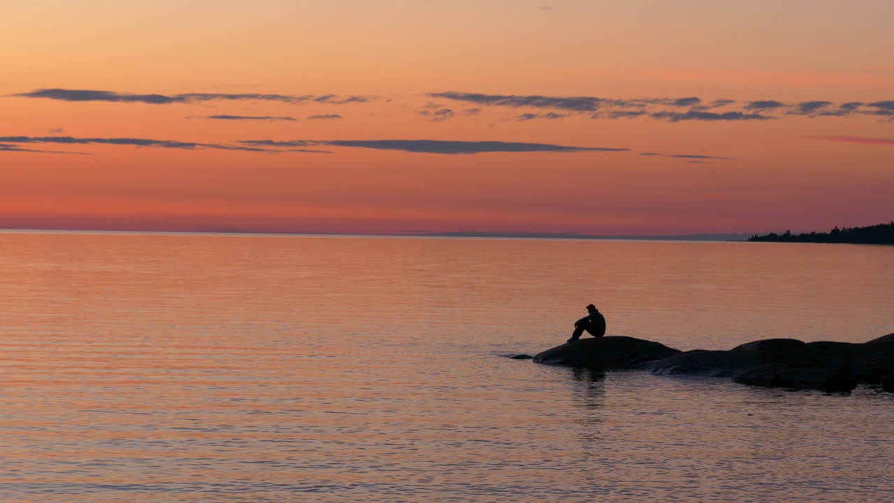 silueta de un hombre en la costa rocosa sentado y disfrutando de la brillante puesta de sol naranja
