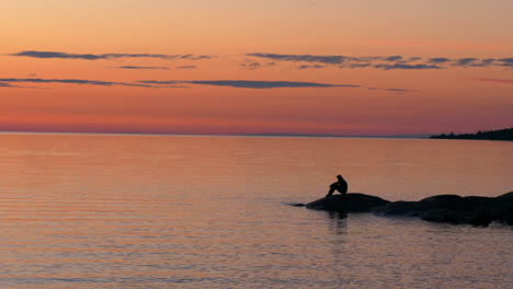 Silhouette-of-a-man-on-rocky-shoreline-sitting-and-enjoying-the-bright-orange-sunset