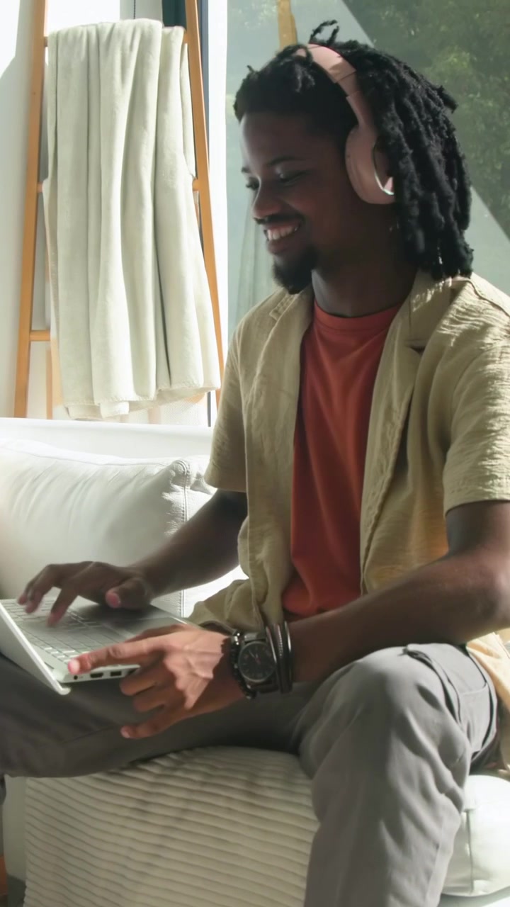 Vertical video: Opening laptop, man typing for work on sofa at home with pink headphones