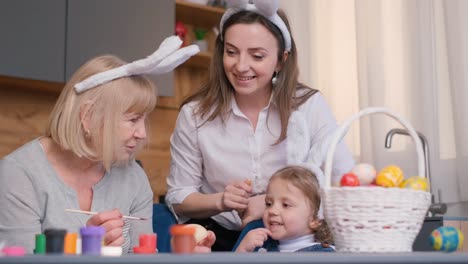 two women and a little girl with bunny ears are happily painting easter eggs at a table. they are sharing this fun activity, capturing the moment with a photograph and smiles on their faces. vertical video