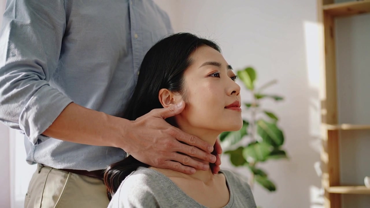 後ろに回り、青いシャツを着た男性が、灰色のトップスを着た座っている女性にカッピングとマッサージを施し、自宅でケアしている様子、植物。