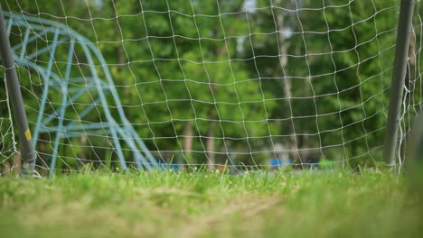 a soccer ball is kicked into a goal post as the goalkeeper misses the ball, on a grassy field with a blur background of outdoor equipment