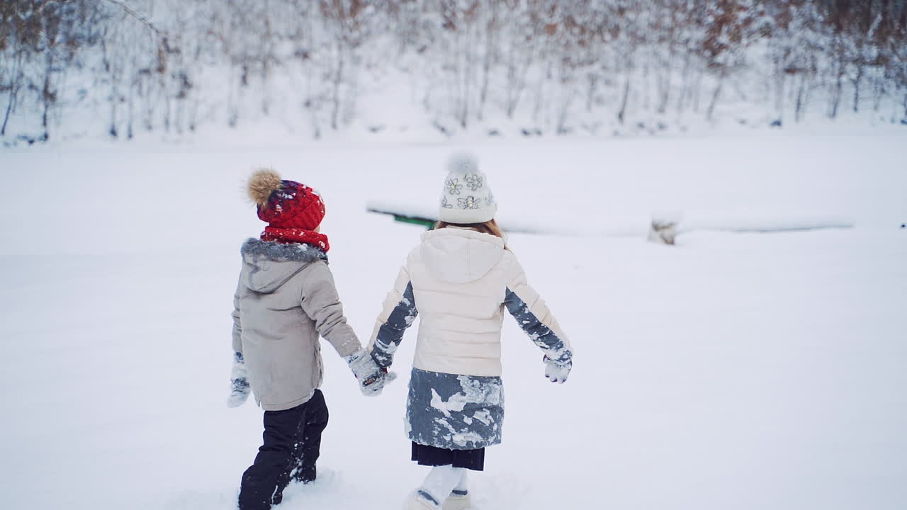 Back view of two children are walking together in the snowy forest. Little kids hold their hands and go through the white snow outdoor. Slow motion.