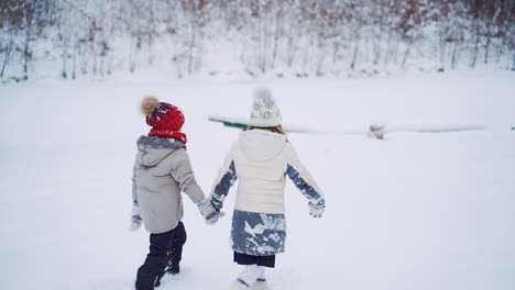 back view of two children are walking together in the snowy forest. little kids hold their hands and go through the white snow outdoor. slow motion.