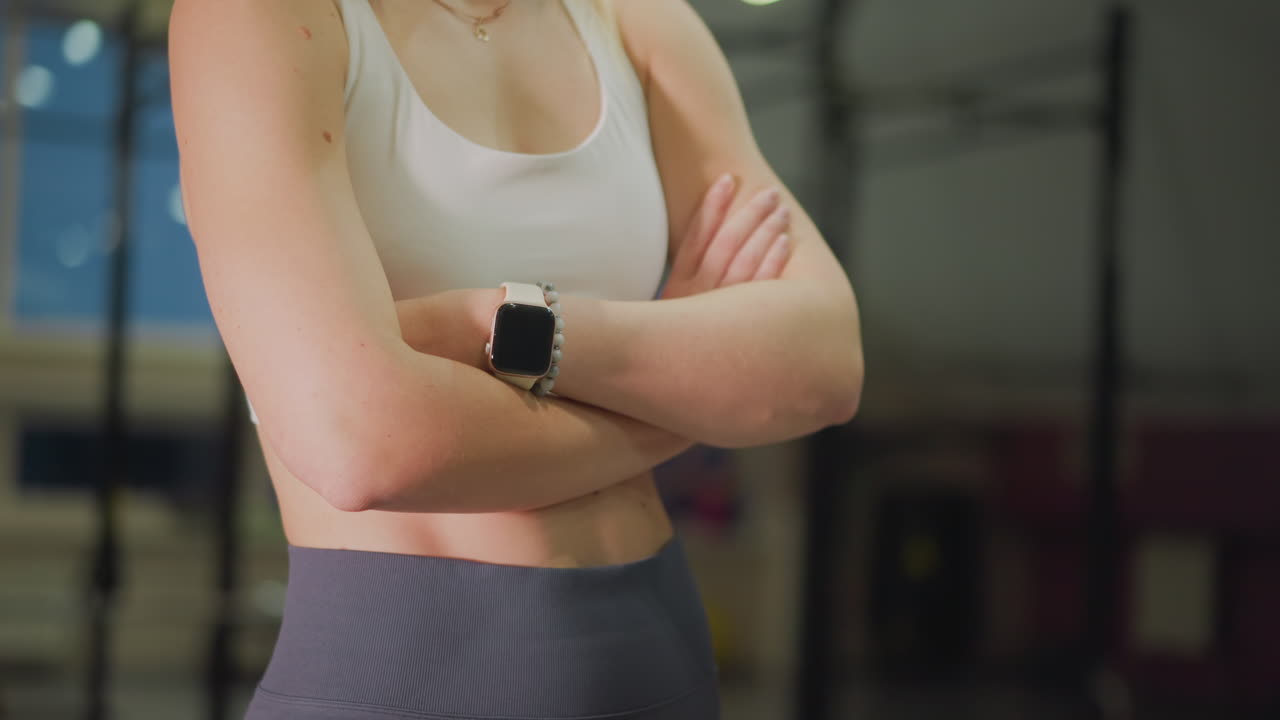 Atleta donna caucasica in piedi a braccia incrociate in palestra, indossa reggiseno sportivo e leggings a vita alta, smartwatch al polso, postura concentrata, illuminazione calda da studio, attrezzature e pesi sullo sfondo, determinazione pre-allenamento.