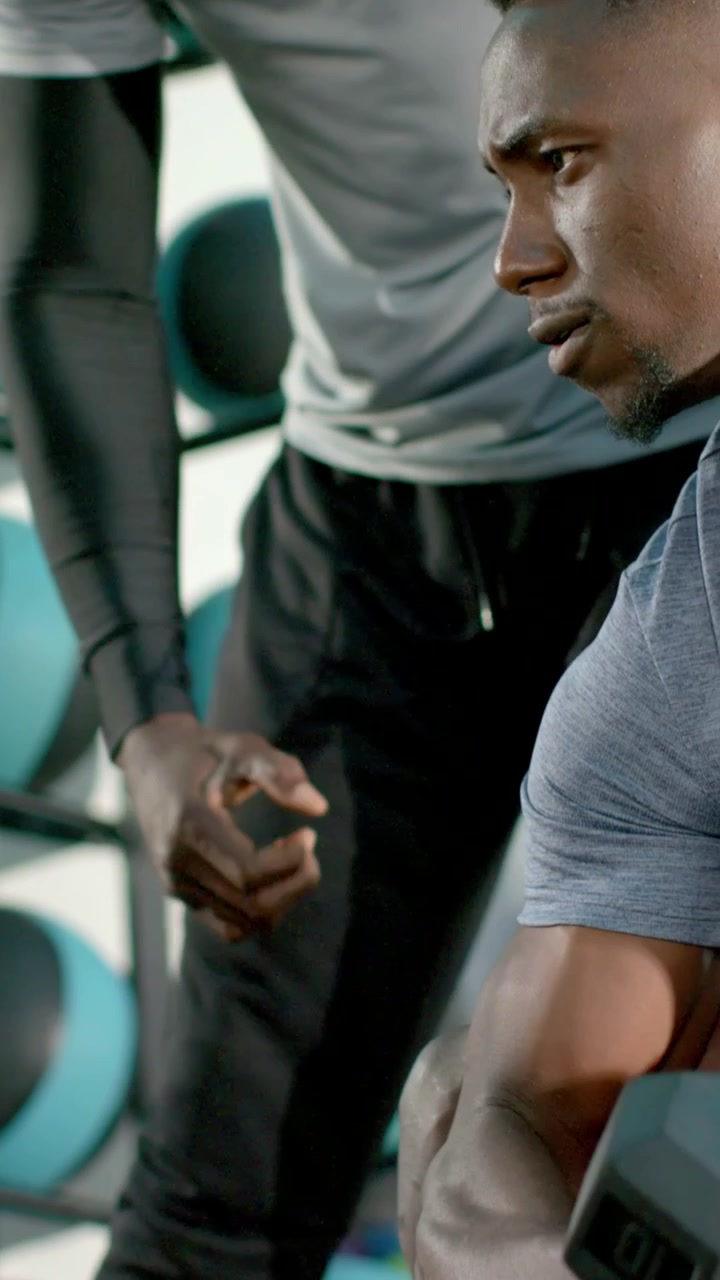 Vertical video: Curling man holding dumbbell at gym as coach guiding form in sleeves, copy space