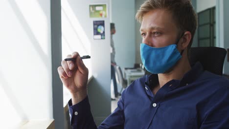 thoughtful caucasian man wearing face mask holding pen sitting on his desk at modern office