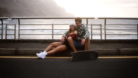 Loving-Sport-Couple-Sitting-On-Border-At-Coastline-Asphalt-Road-On-Background-Hills-And-Foggy-Ocean