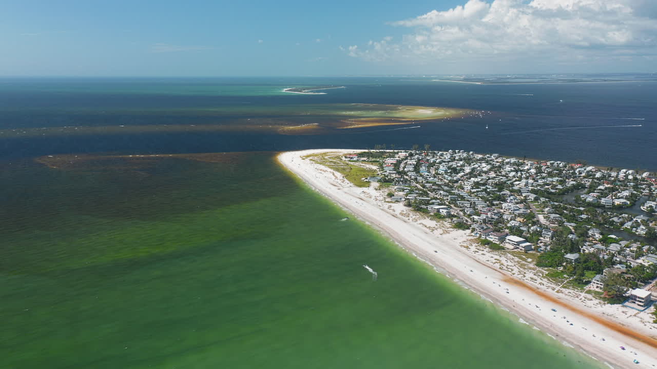 Turquoise waters meet the sandy beaches of Anna Maria Island as residential neighborhoods line the coastline, with distant sandbars and ocean horizon visible
